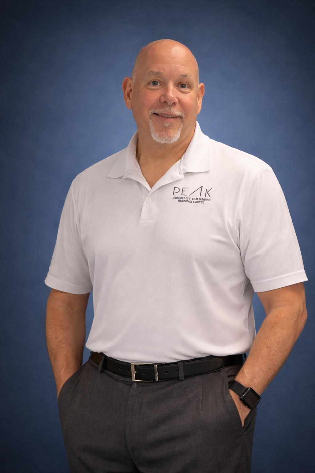 Senior Instructor Scott Walters. A smiling middle-aged man with a bald head and goatee, wearing a white polo shirt with a logo and text, dark trousers, and a smartwatch, standing against a dark blue background.