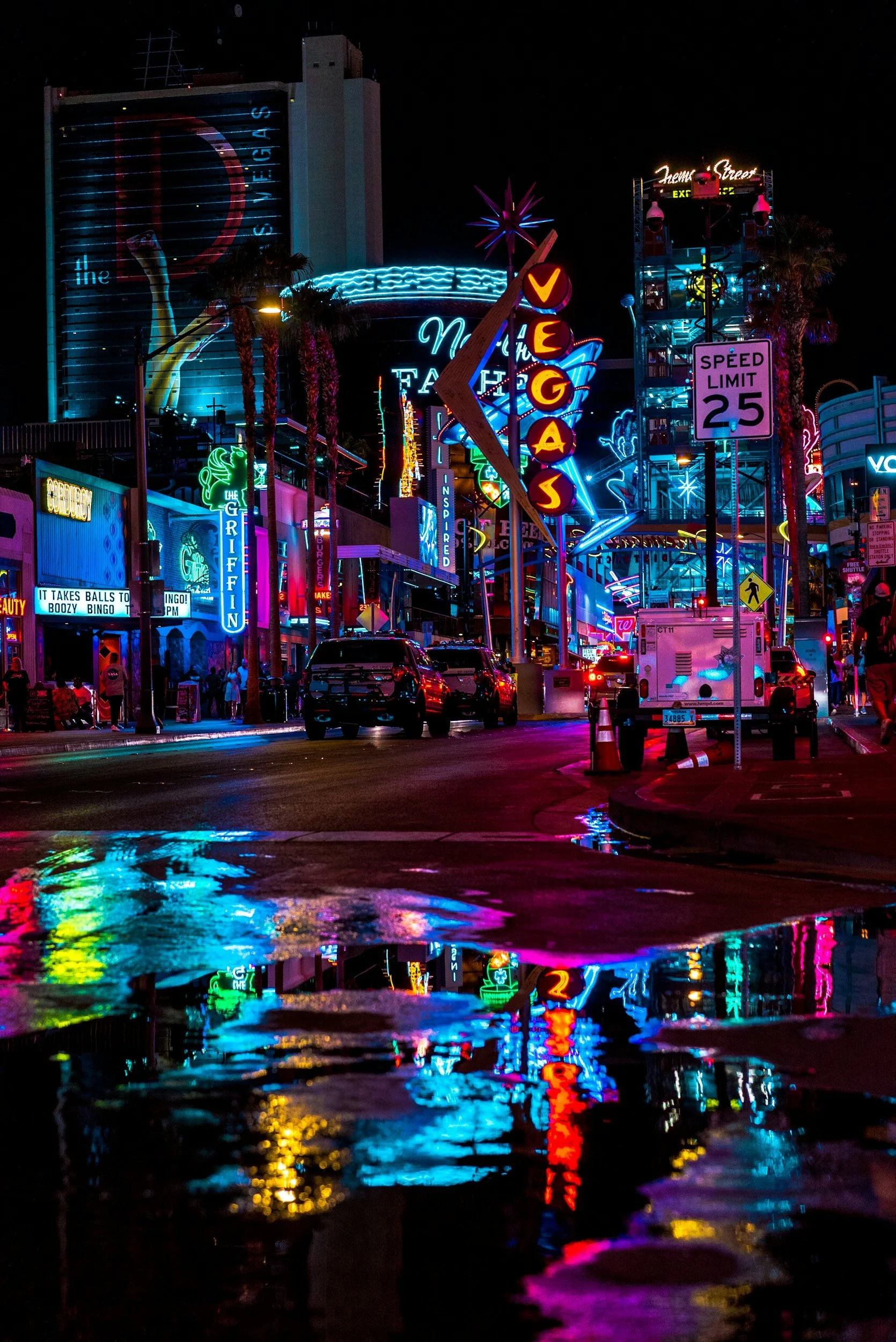 Night view of a neon-lit street with colorful signs, including a large 'Vegas' sign, and reflections on wet pavement.