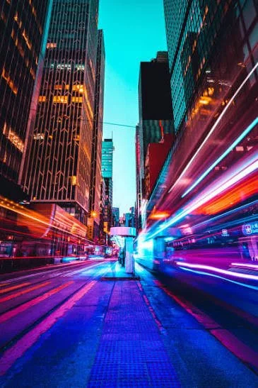 A vibrant city street at night with tall glass skyscrapers on both sides and colorful light streaks from moving vehicles.