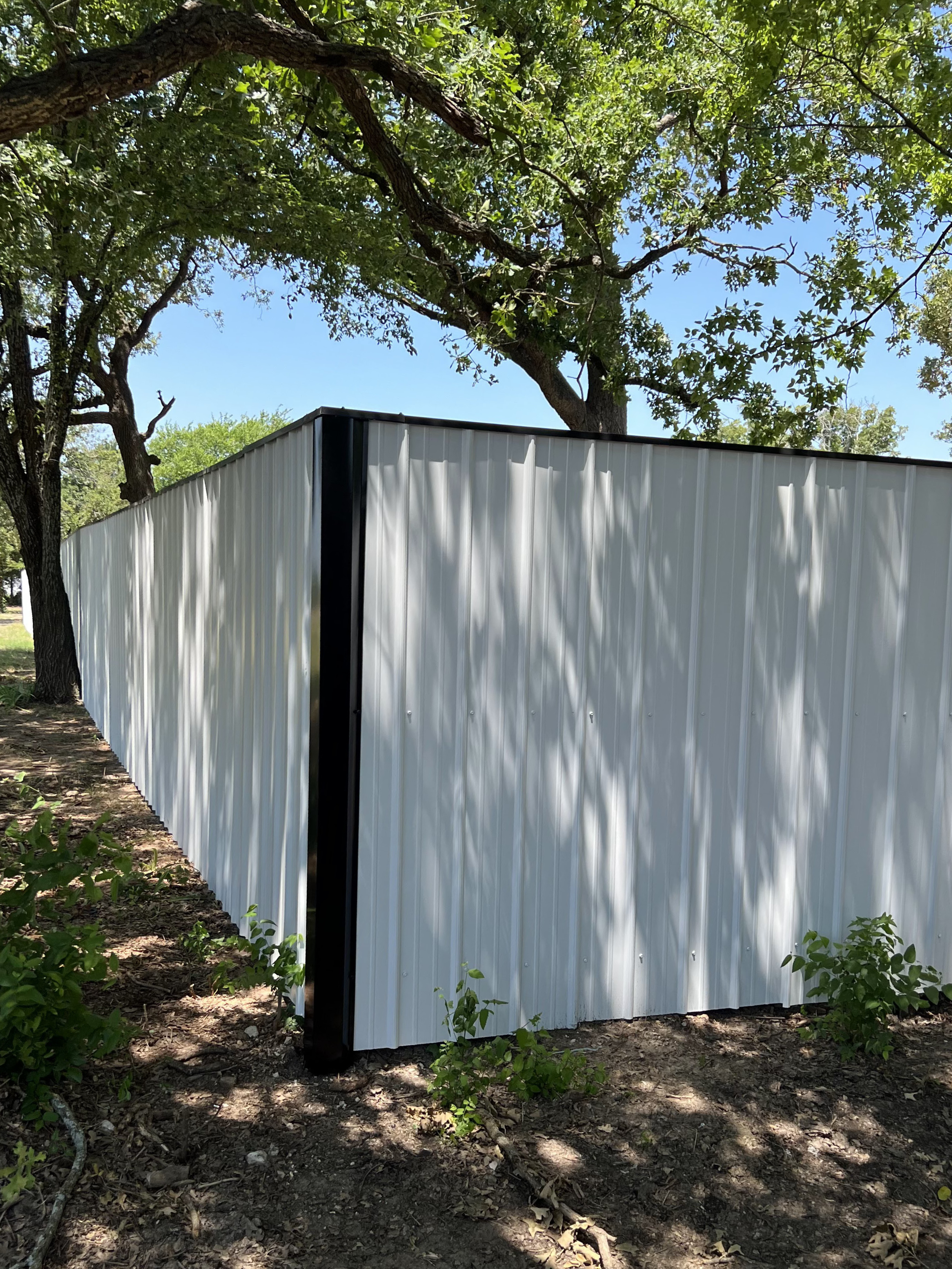 A white metal fence with vertical ridges along a dirt path with green plants, shade from trees, and a clear blue sky.
