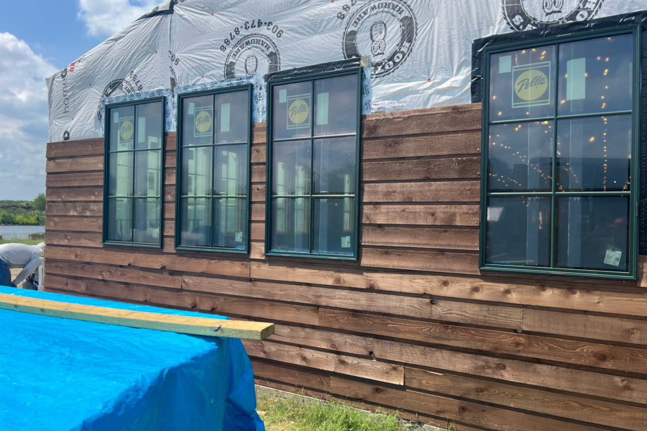 Building under construction with dark green trimmed windows, brown wooden siding, and white weather-resistant wrap on top.