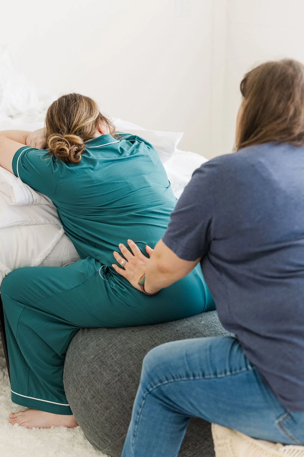 A woman in teal sitting on an exercise ball beside a hospital bed, resting her head on a pillow, while another woman in a gray shirt is sitting beside her, holding her hand and providing comfort.