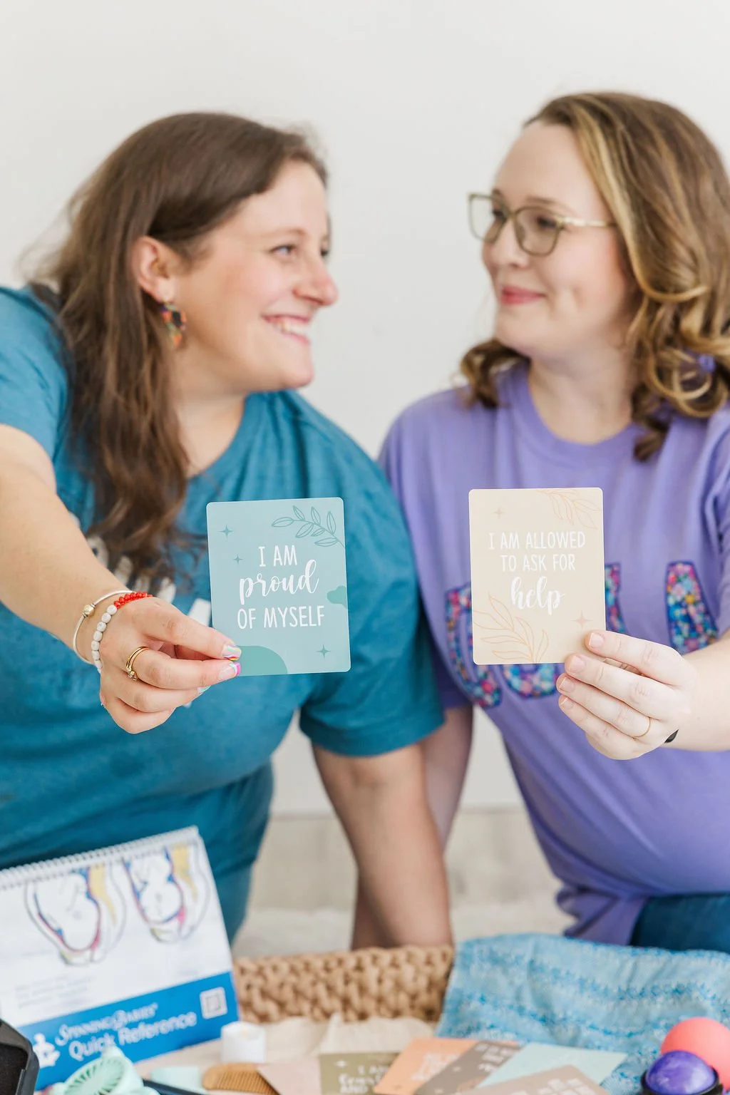Two women smiling and holding up cards with positive affirmations. The woman on the left has brown hair and wears a teal shirt, while the woman on the right has curly hair, glasses, and wears a purple shirt.