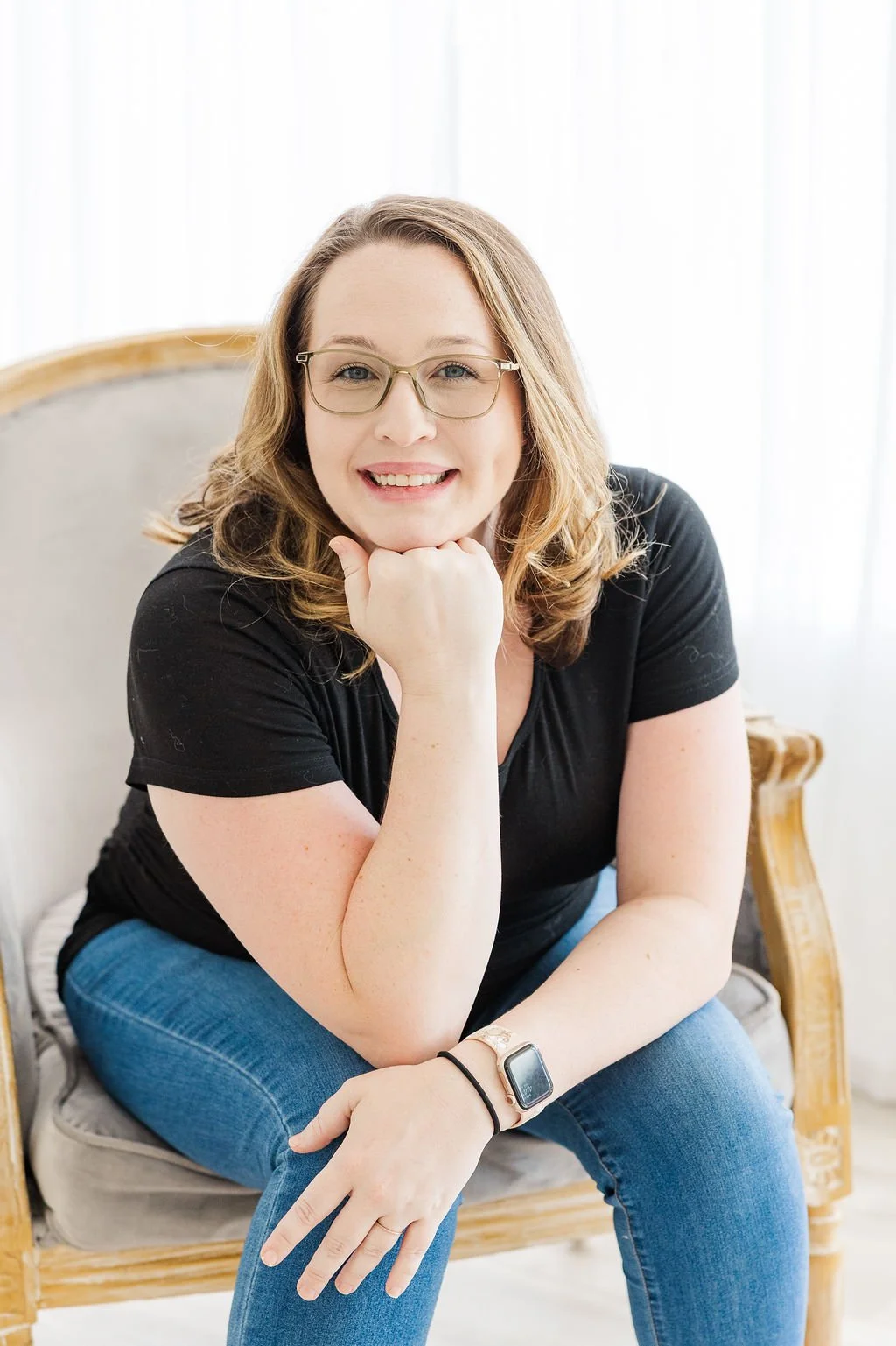 Portrait of a woman with shoulder-length blonde hair and glasses, smiling and resting her chin on her hand, sitting on a vintage-style chair in a bright room.