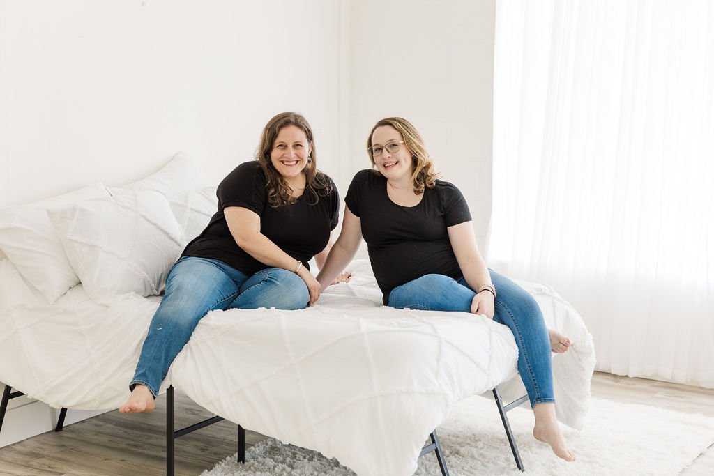 Two women sitting on a bed, smiling at the camera, in a bright room with white curtains.