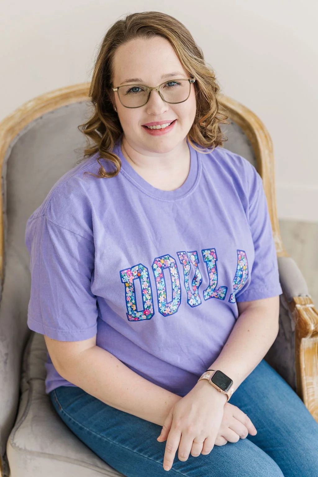 A woman with glasses and red hair sitting on a vintage-style chair, wearing a purple T-shirt with floral lettering that says 'DOULA' and jeans.
