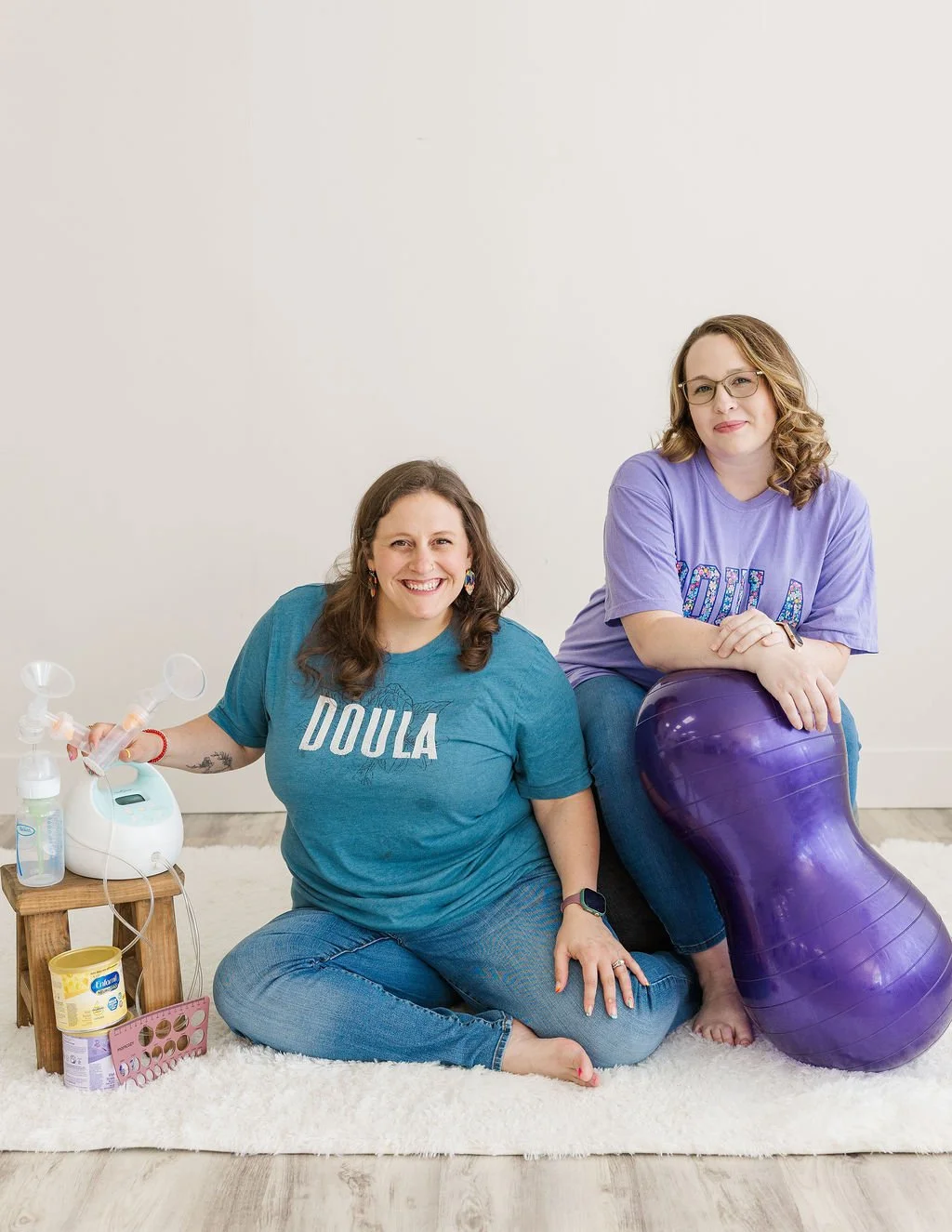 Two women wearing Doula shirts sitting on a rug against a plain wall. One woman smiling, sitting on the floor with a breast pump nearby, others with a purple birthing ball.