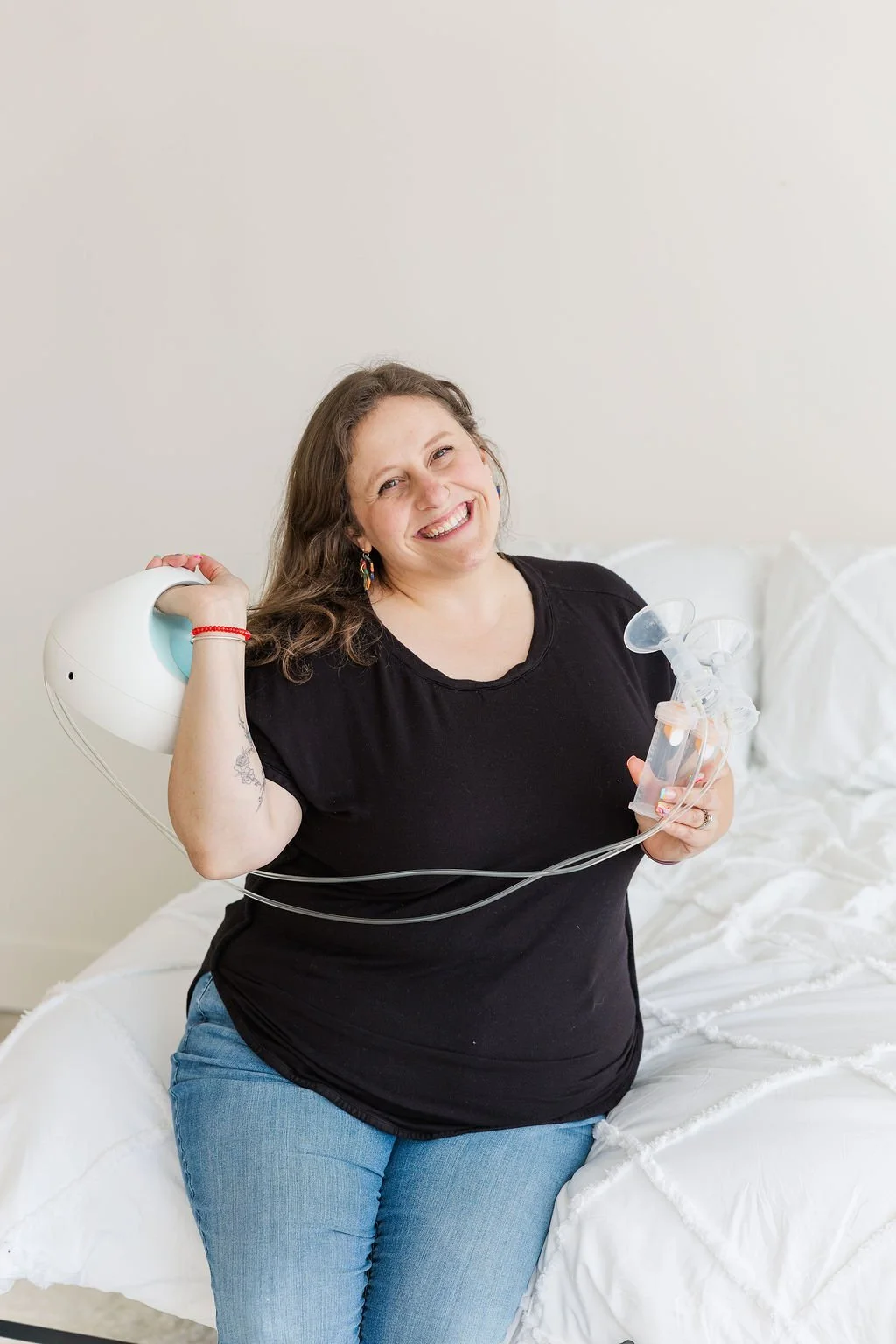 A smiling woman holding a breast pump and a bottle on a bed with white bedding.