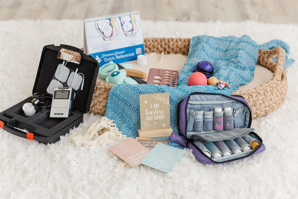 A collection of self-care and wellness items including a TENS unit in a black case, essential oils, a spinning babies book, a massage roller, and a blue Rebozo on a white fluffy surface.
