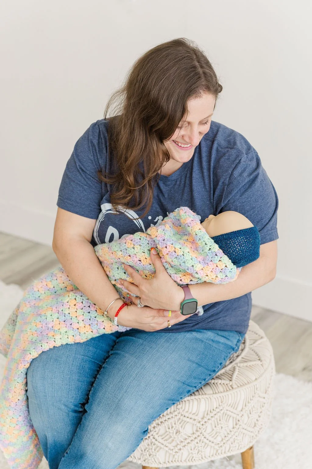 A woman with brown hair, wearing a blue t-shirt and jeans, smiles while holding a baby wrapped in a colorful crocheted blanket and wearing a blue knit cap.