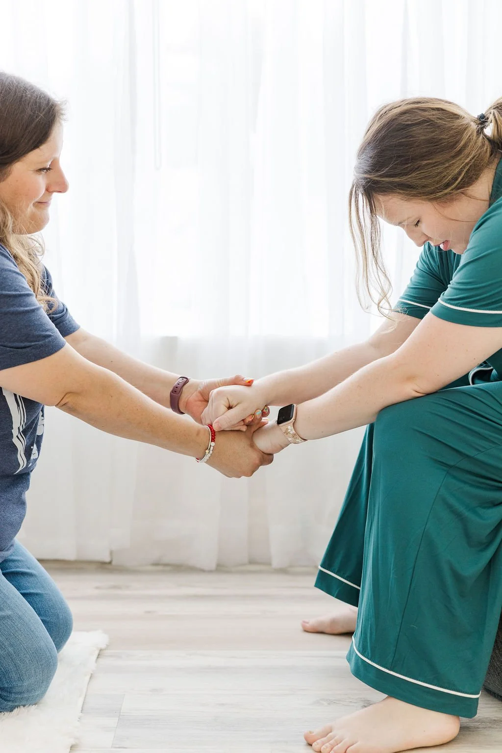 Two women holding hands and gripping each other's wrists in a supportive stance indoors, with white curtains in the background.
