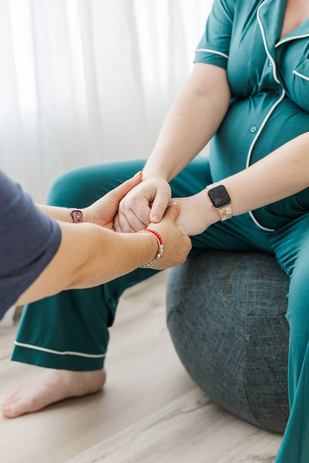 A person with a smartwatch on their wrist sitting on a gray exercise ball, holding hands with another person who is kneeling on the floor offering support.