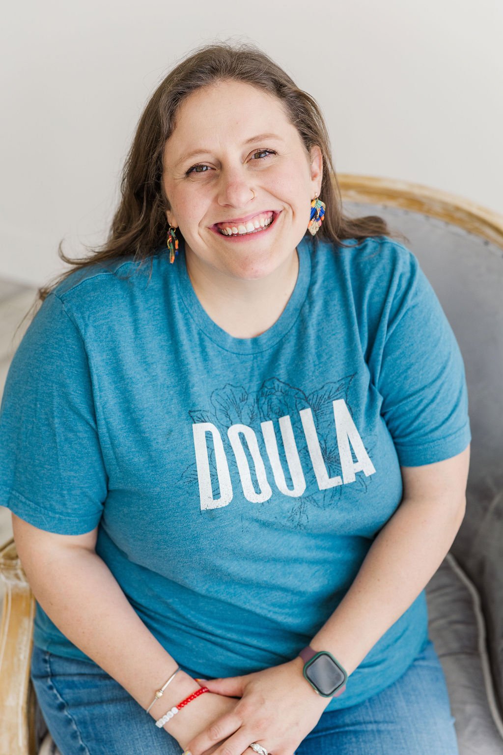 A smiling woman with curly brown hair, wearing a blue t-shirt that says "DOULA," colorful earrings, and a smartwatch, sitting on a chair with a light-colored wall in the background.