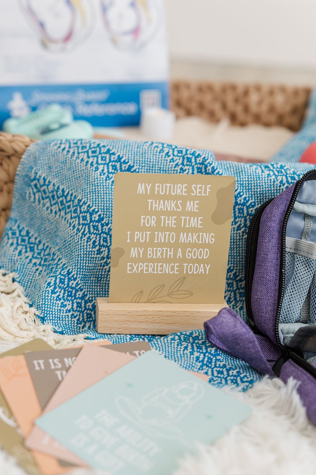 A framed sign with the quote 'My future self thanks me for the time I put into making my birth a good experience today,' surrounded by baby items including a purple and gray diaper bag, a blue patterned cloth, and colorful cards, with a blurry background of baby supplies.