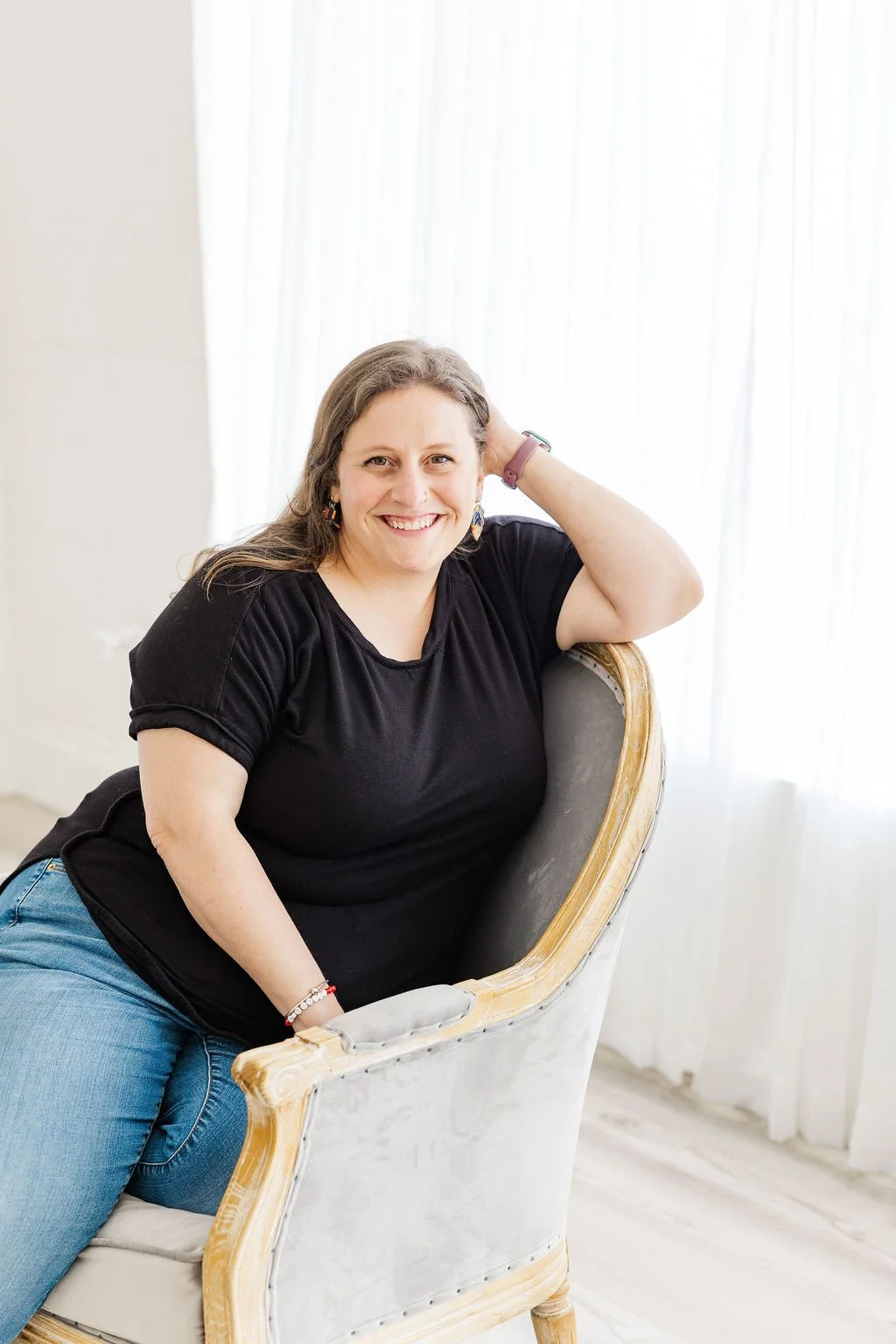 Woman with long brown hair, wearing a black t-shirt and blue jeans, smiling and sitting on a vintage armchair with a beige upholstery, in front of white curtains.