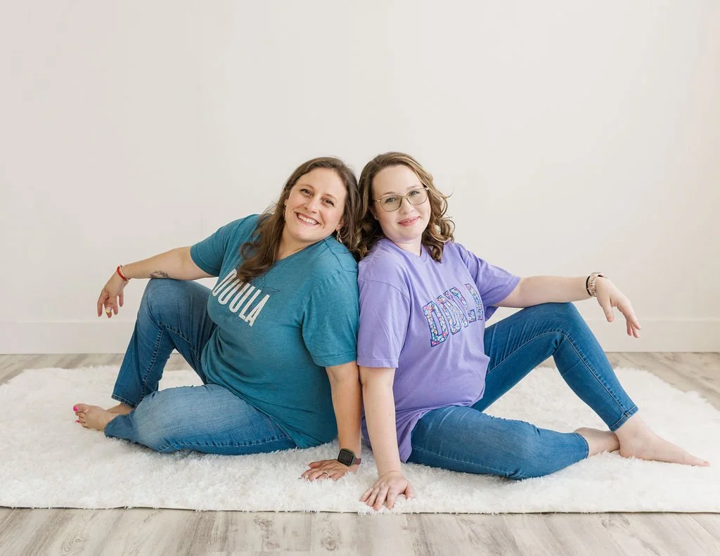 Two women sitting on a white rug back-to-back, smiling at the camera, in a room with white walls and wooden floor.