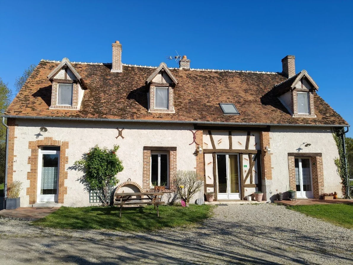 Maison traditionnelle avec un toit en tuiles, fenêtres à volets, jardin et un chemin en gravier devant.