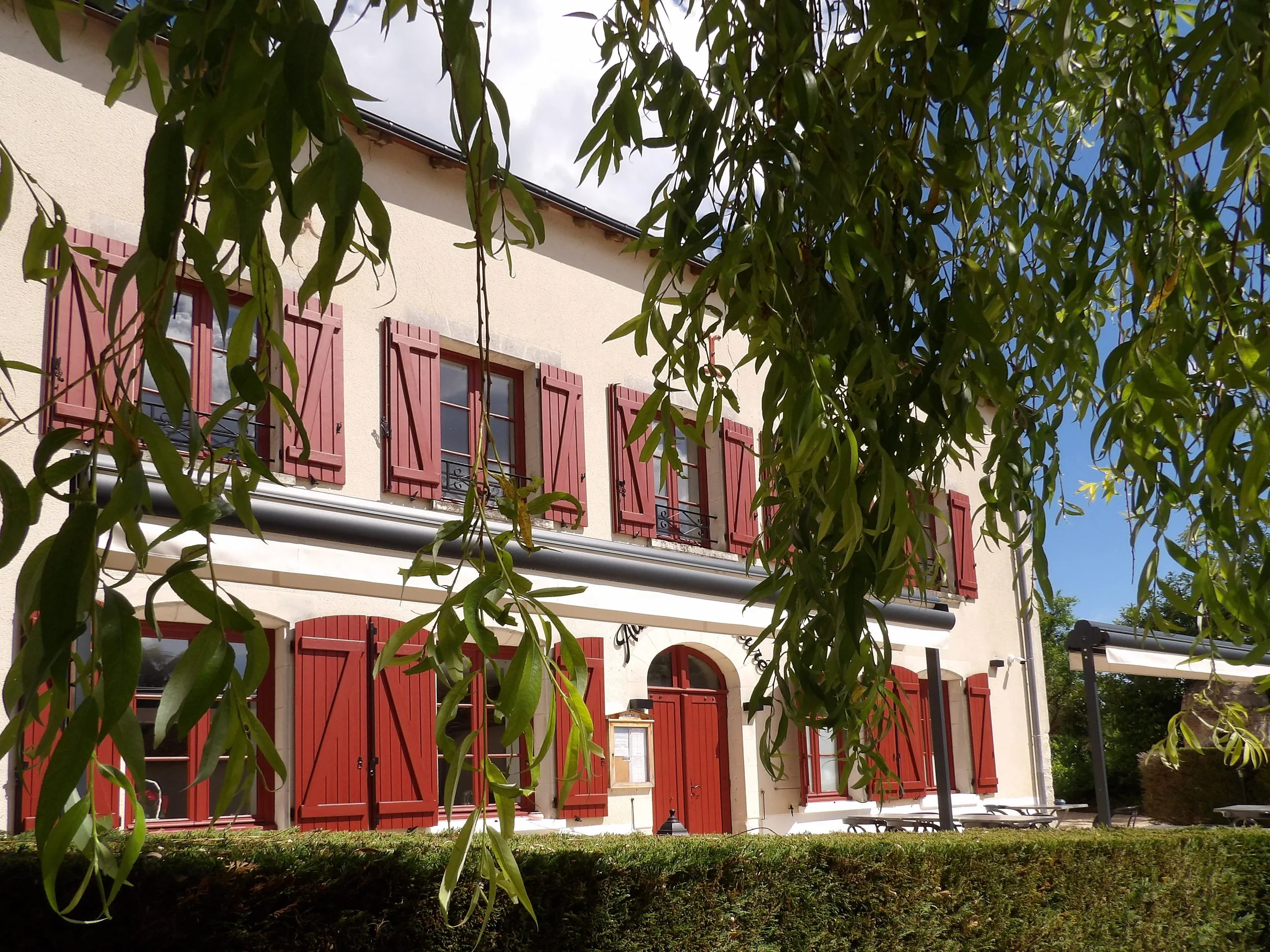 Une maison avec des volets rouges, entourée d'arbres et de verdure, sous un ciel partiellement nuageux.