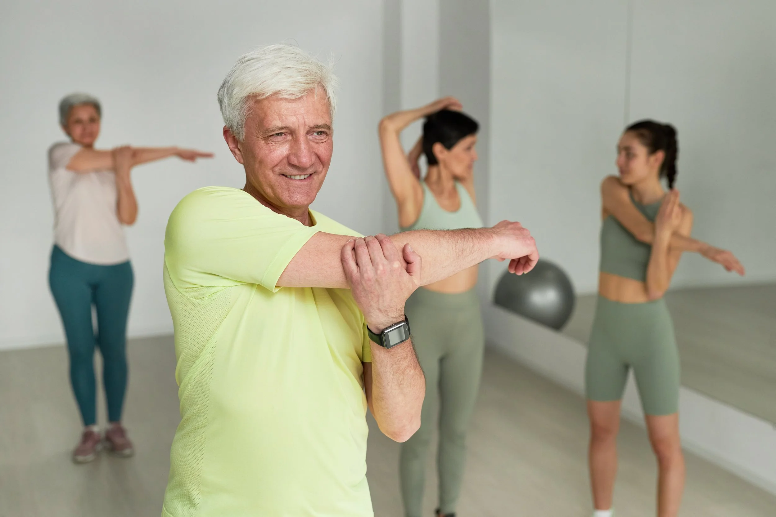 Un homme âgé en tenue de sport réalise un étirement de bras lors d'une séance de fitness avec trois femmes en arrière-plan dans une salle de sport.