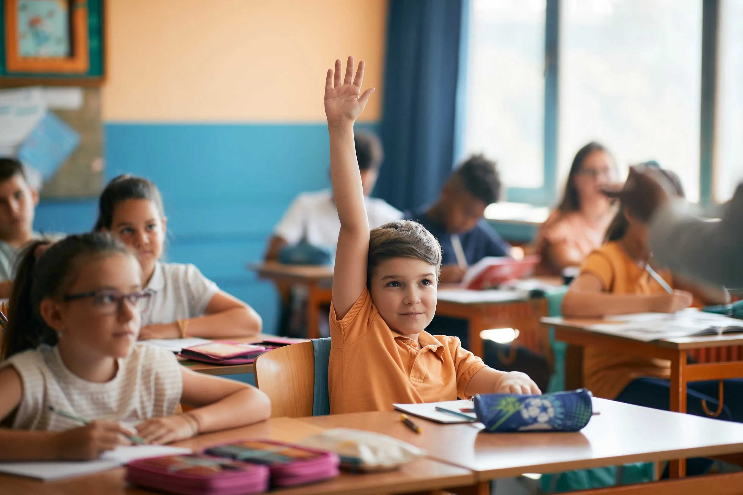 Une classe avec des enfants, un garçon en orange lève la main, plusieurs autres élèves assis, une fenêtre laissant entrer la lumière.
