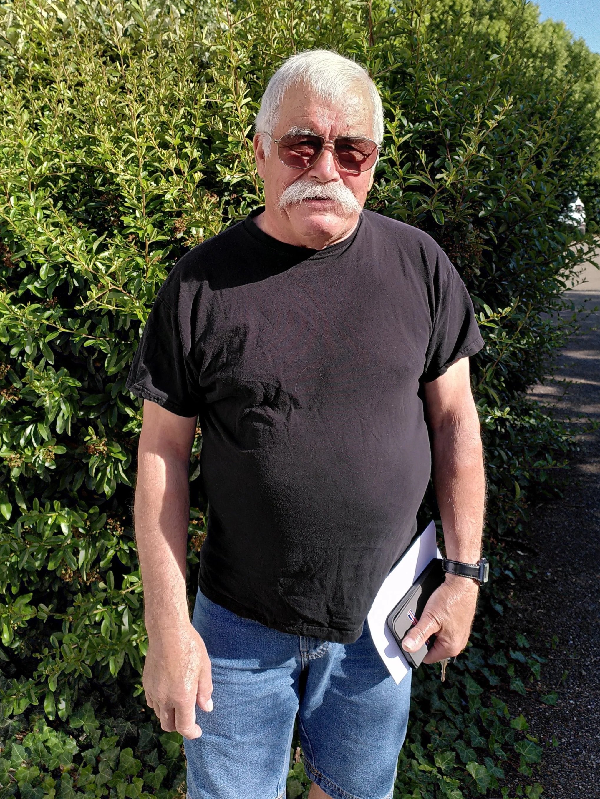Un homme âgé avec des cheveux gris, une moustache et une barbe blanche, portant des lunettes de soleil marron, en t-shirt noir, tenant un téléphone et un carnet blanc, debout devant un buisson vert sous un ciel bleu.