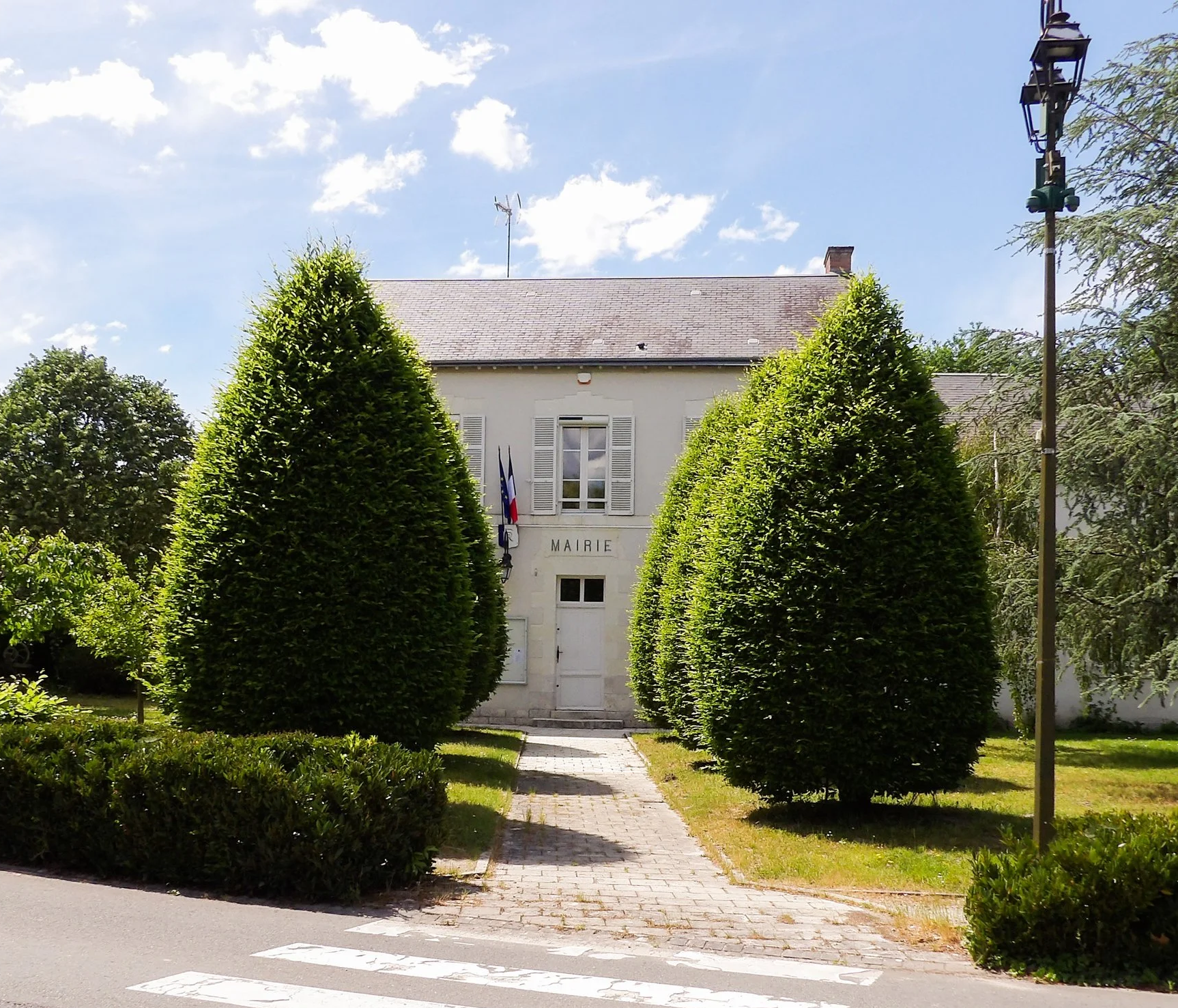 Bâtiment de la mairie français avec des drapeaux, entouré d'arbustes et d'arbres, sous un ciel en partie nuageux.