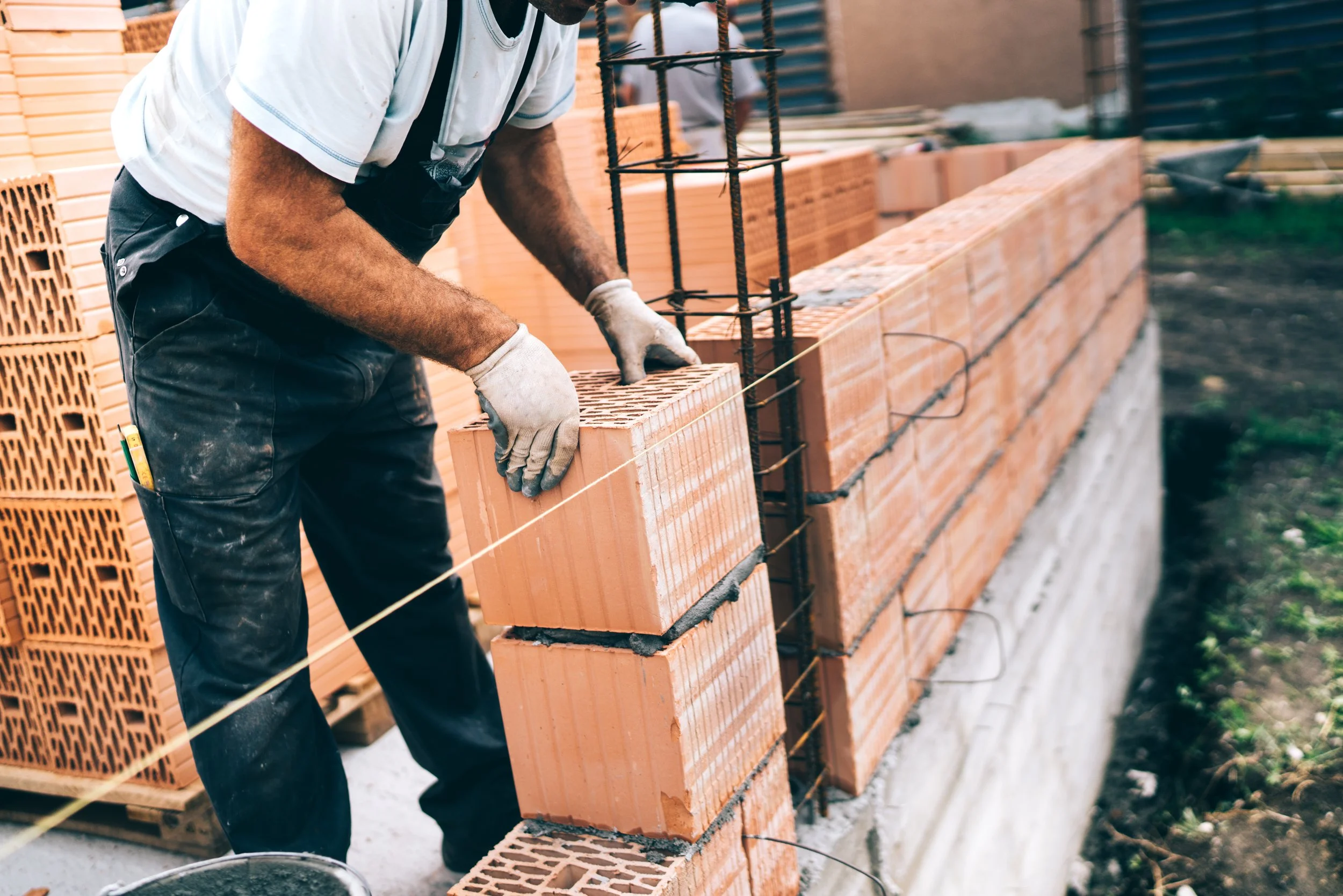 Un homme construit un mur en briques sur un chantier, portant des gants et une t-shirt blanc, avec des rangées de briques orange à l'arrière-plan.