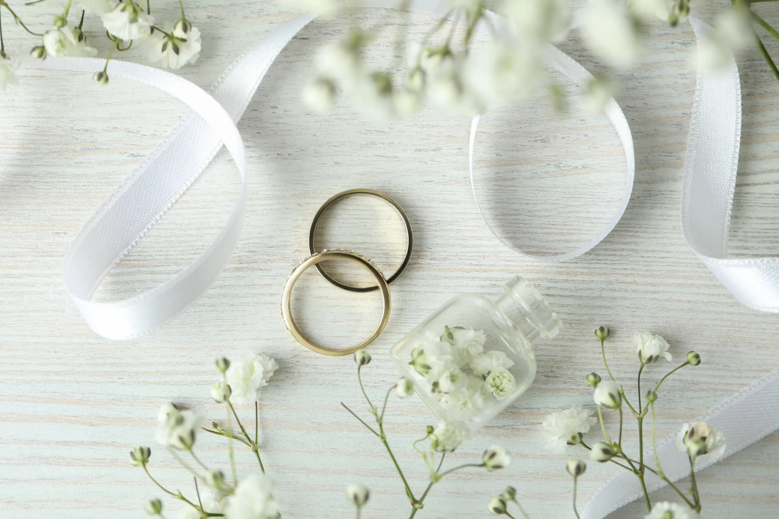 Anneau de mariage doré et anneau en métal argenté posés sur une surface en bois clair, entourés de ruban blanc, petites fleurs blanches et petits vases avec des fleurs, décoration délicate et élégante pour un mariage.