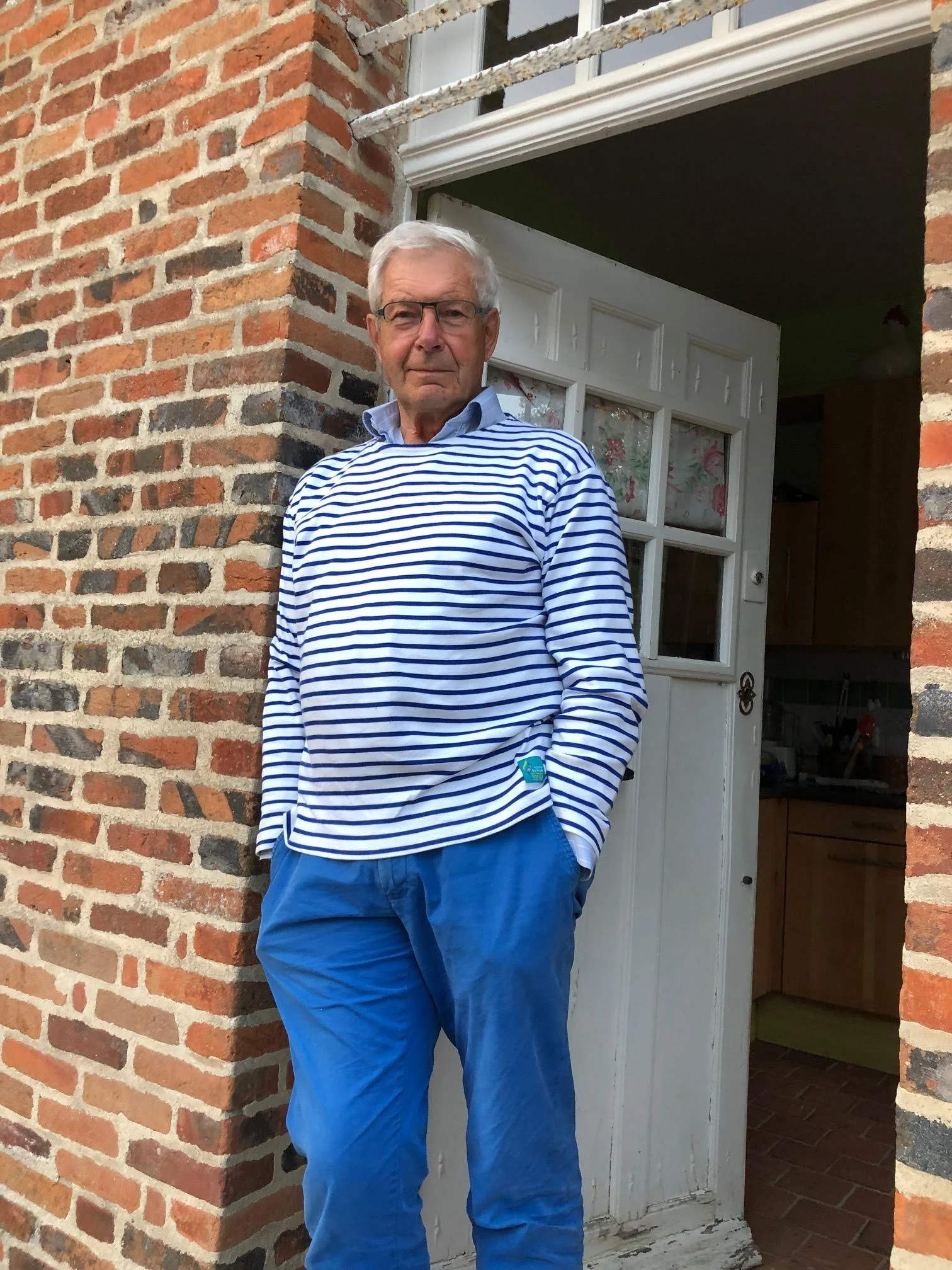 Un homme âgé à cheveux blancs porte un chemisier à rayures bleues et blanches et un pantalon bleu, se tenant près d'une porte blanche avec un mur en briques rouge. Il porte des lunettes et regarde vers l'appareil photo.