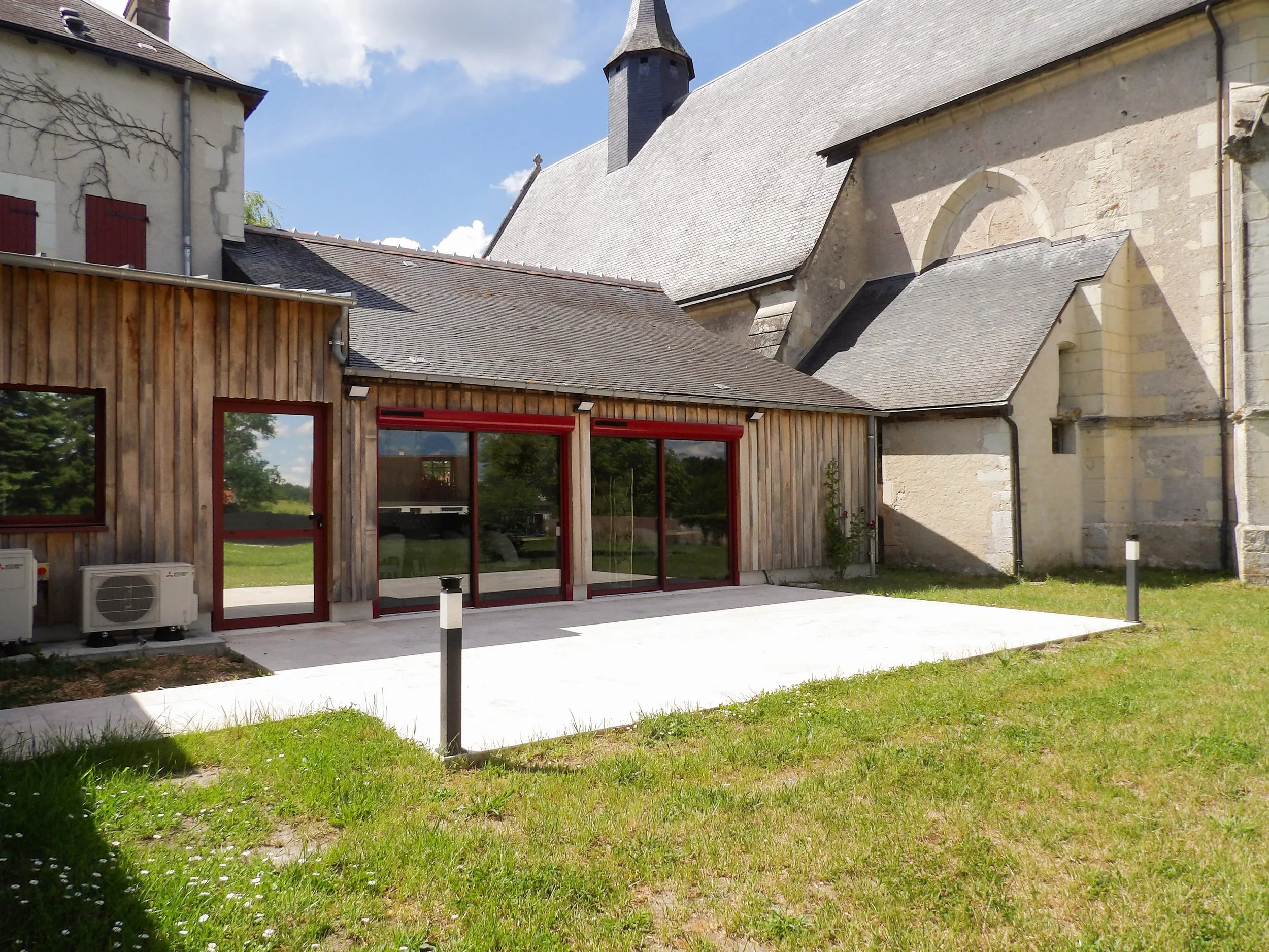 Vue extérieure d'une ancienne église avec un toit en ardoise, une structure en pierre et une partie moderne en bois avec fenêtres à cadre rouge, entourée d'une pelouse verte et un ciel bleu avec quelques nuages.