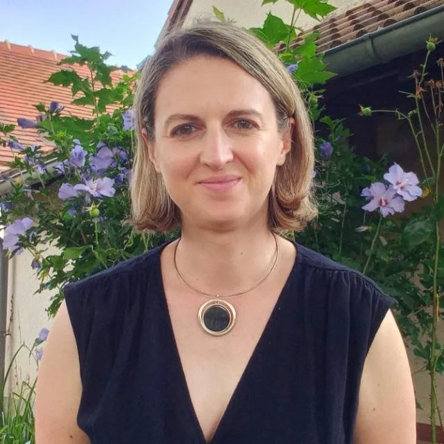 Une femme avec des cheveux blonds moyens, portant un haut noir sans manches et un collier rond, se tenant devant des fleurs violettes et des feuilles vertes dans un jardin, sous un ciel bleu.