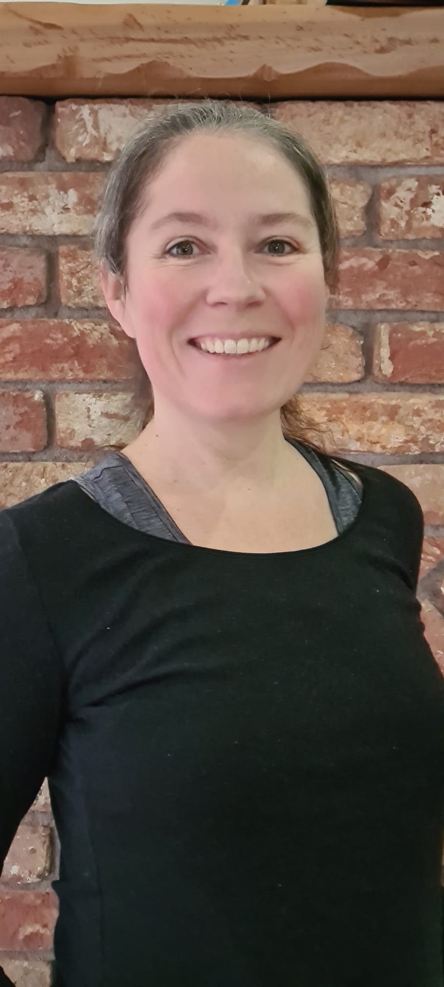 Une femme souriante avec des cheveux bruns attachés, portant un haut noir avec des détails gris aux épaules, se tenant devant un mur en briques.