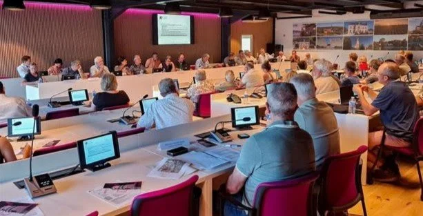Une salle de conférence avec des participants assis à des bureaux, regardant un écran de présentation à l'avant.