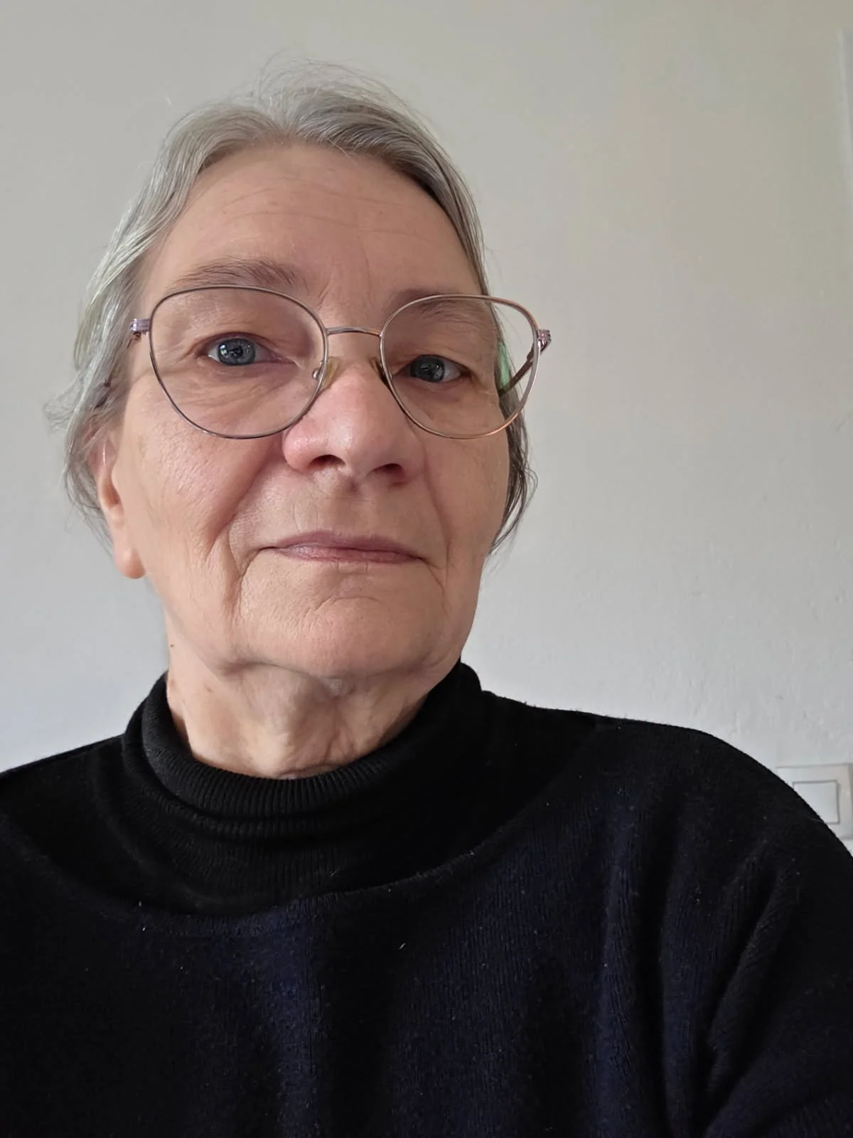 Portrait d'une femme âgée portant des lunettes et un pull noir, avec un fond beige clair.