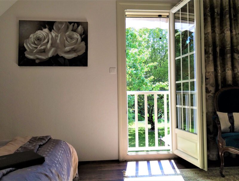 Une pièce avec un balcon ouvert laissant voir un arbre vert à l'extérieur, un lit partiellement visible avec une couverture gris clair, un tableau de fleurs blanches en noir et blanc, et un fauteuil ancien en bois avec un coussin.