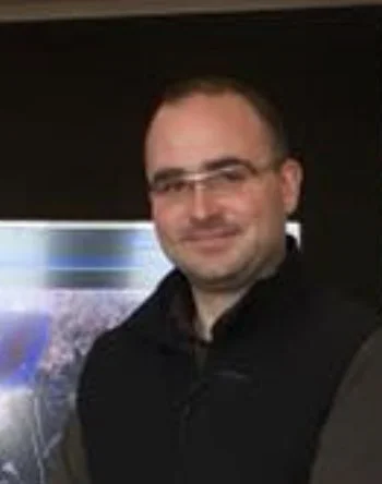 Homme souriant portant des lunettes, fond sombre.