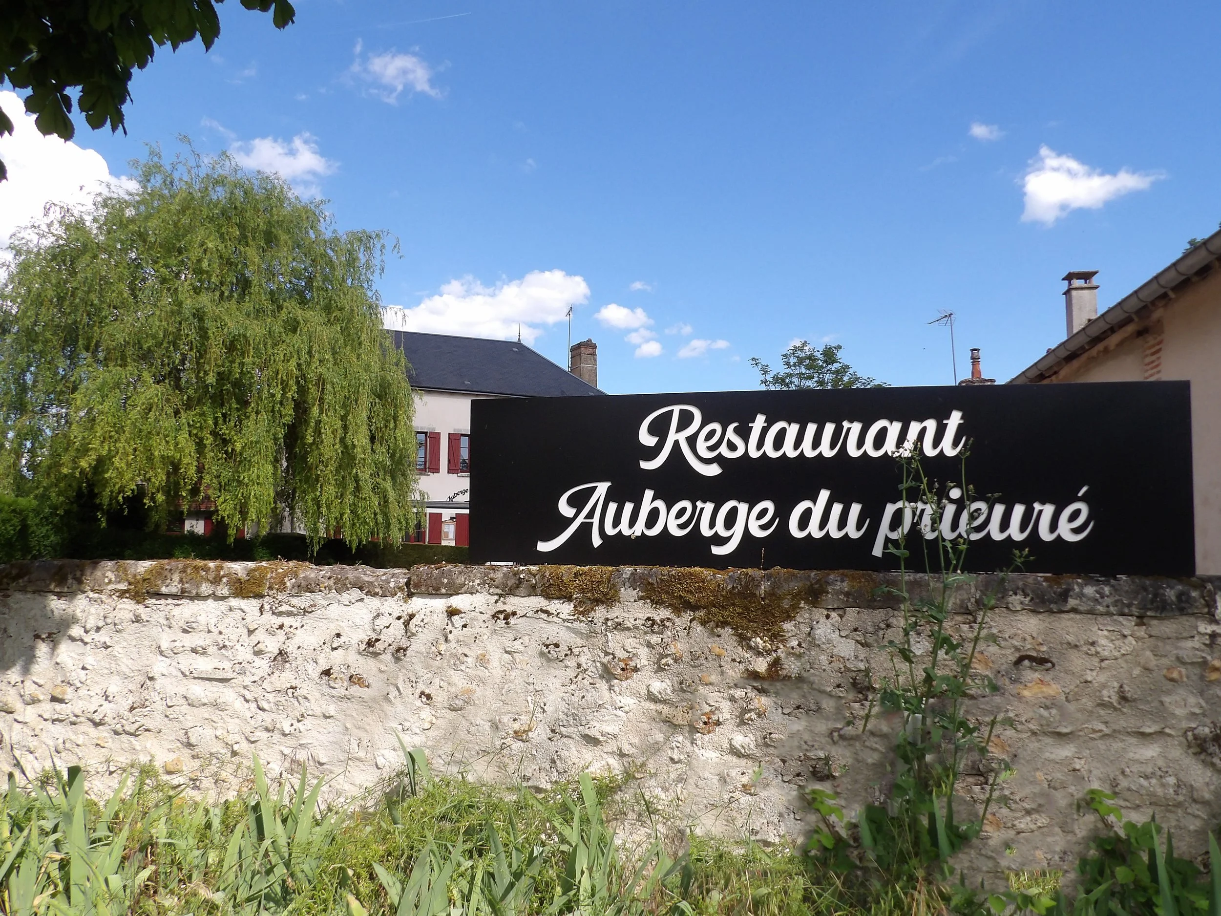 Une enseigne indiquant un restaurant et une auberge du peintre devant un mur en pierre. En arrière-plan, des maisons avec des toits inclinés, des cheminées et des antennes parabolique, sous un ciel bleu clair avec quelques nuages.