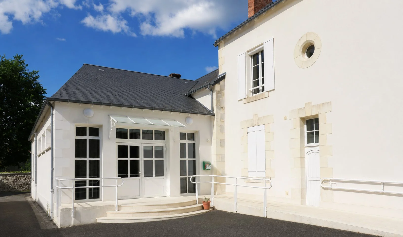 Façade blanche d'une maison avec un sapin en fond et un ciel bleu avec des nuages.