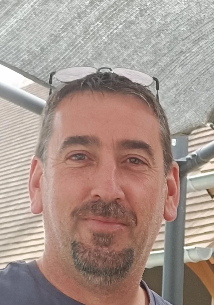 Un homme souriant avec des lunettes sur le dessus de la tête, sous un parasol, devant un bâtiment en bois.