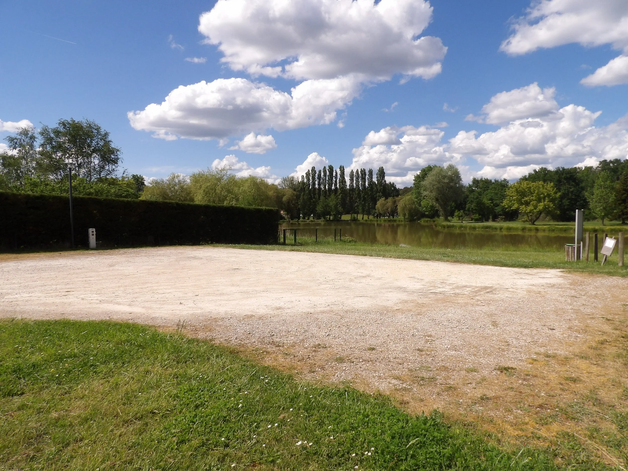 Un paysage de parc avec un lac, des arbres feuillus et un ciel bleu avec des nuages blancs.