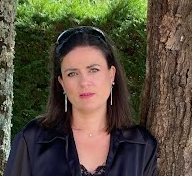 Une femme avec des cheveux noirs et des yeux bleus, portant une veste noire, debout près d'un arbre dans un environnement extérieur verdoyant.