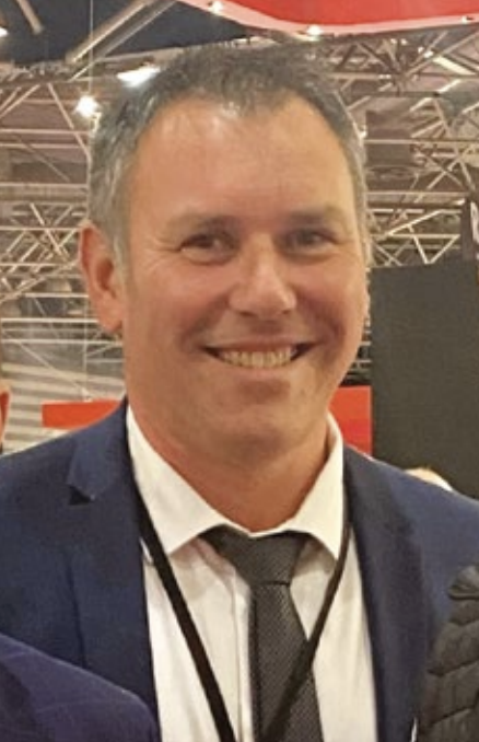 Un homme souriant en costume, portant une chemise blanche, une cravate grise et un badge, dans un environnement intérieur avec structures métalliques et éclairage.
