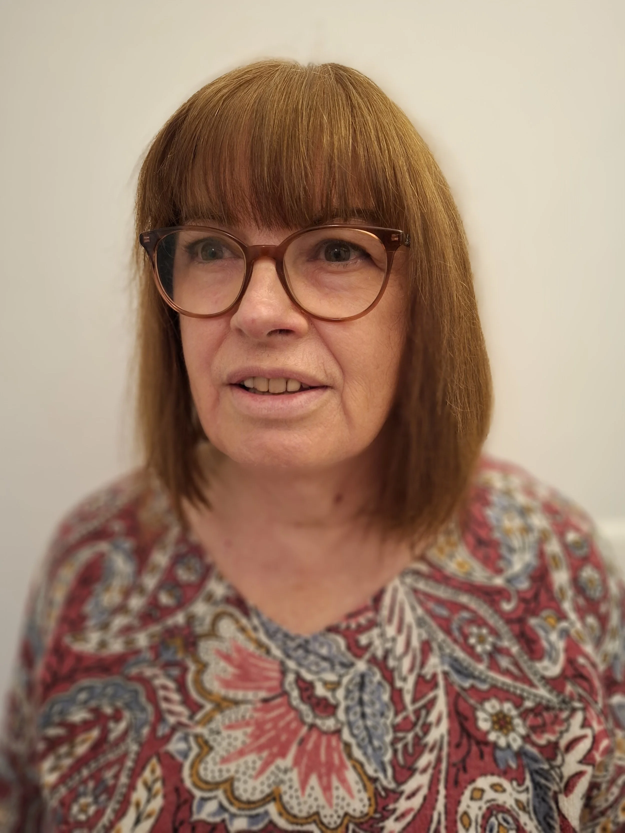 Une femme avec des cheveux roux, portant des lunettes carrées, souriant légèrement, portant un vêtement à motif floral coloré, debout contre un mur blanc.