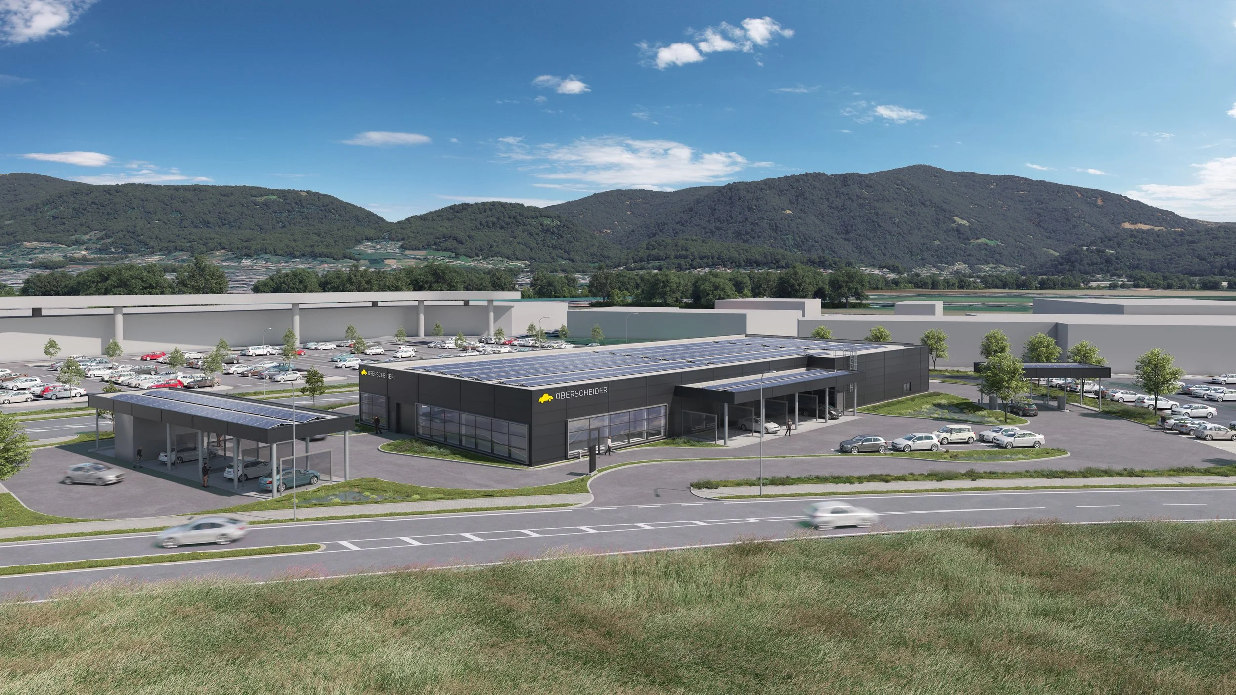 Neues Autohandelszentrum namens Oberscheider mit Solarzellen auf dem Dach, umgeben von Parkplätzen und einer Straße, mit Bergen im Hintergrund und blauem Himmel mit wenigen Wolken.