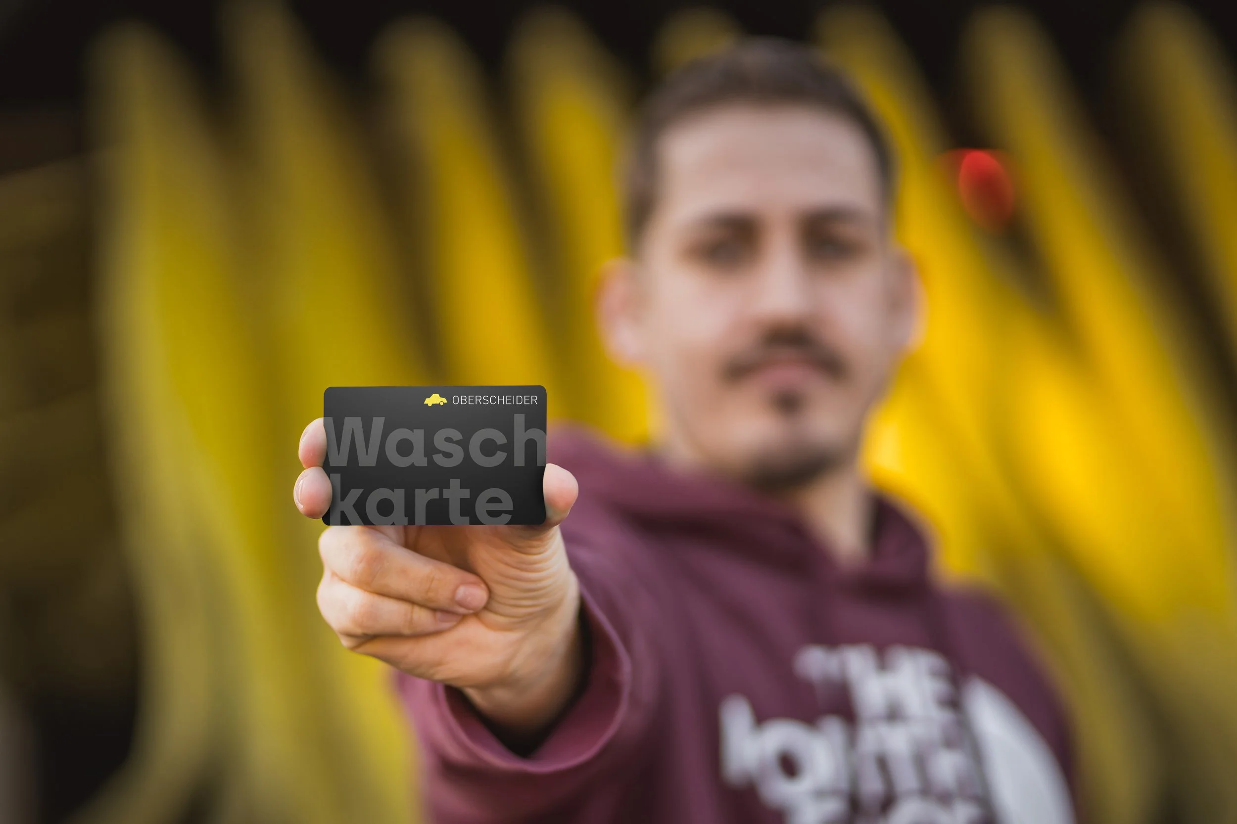 Ein Mann hält eine schwarze Karte mit weißen Buchstaben, auf der steht "Waschkarte", während er vor einem gelben Hintergrund steht.