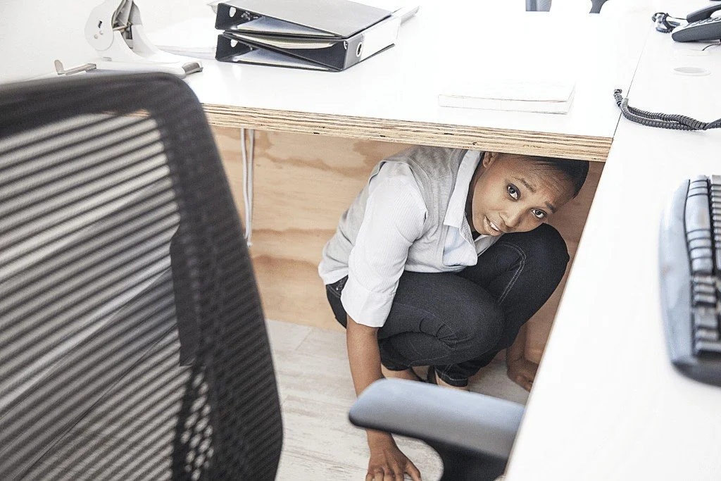 A woman crouching under a white office desk, looking upward with a surprised expression. Workplace Safety. Active Threat.