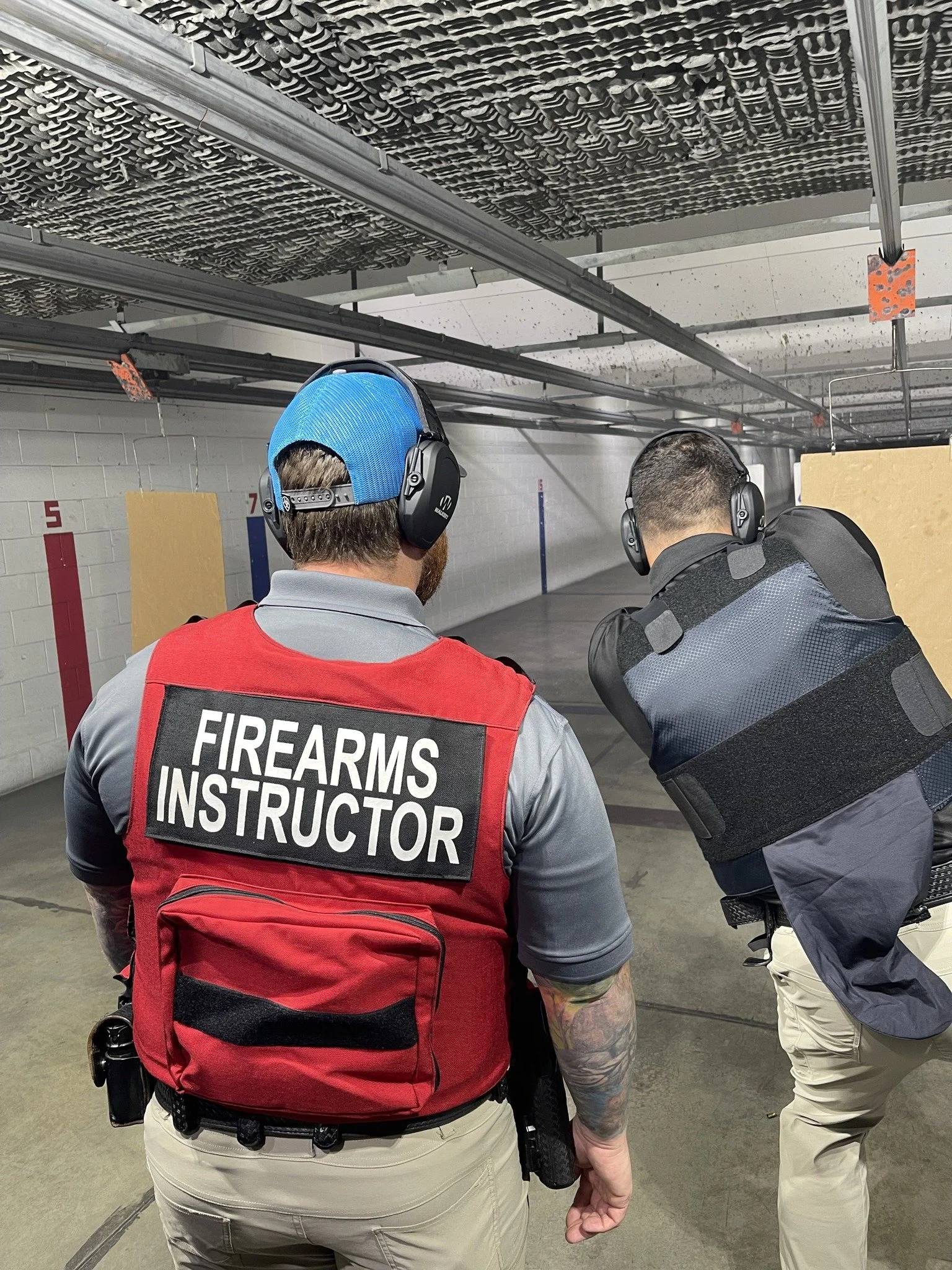 Two firearms instructors at an indoor shooting range, wearing headsets, one with a red vest labeled 'FIREARMS INSTRUCTOR,' and the other in a black tactical vest, are standing side by side, facing the shooting range.