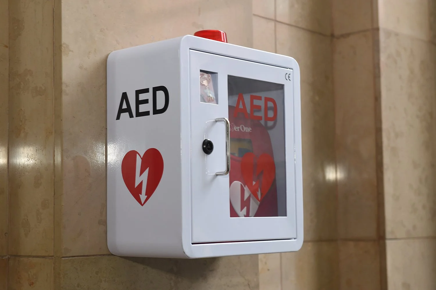 A white AED (automated external defibrillator) cabinet mounted on a beige tiled wall, with a glass door displaying a red heart with a white lightning bolt, and the letters 'AED' printed on it.