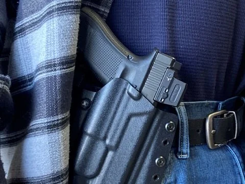 A person wearing a plaid shirt with a black pistol holstered on their waistband. Permit to Carry. Renewal.