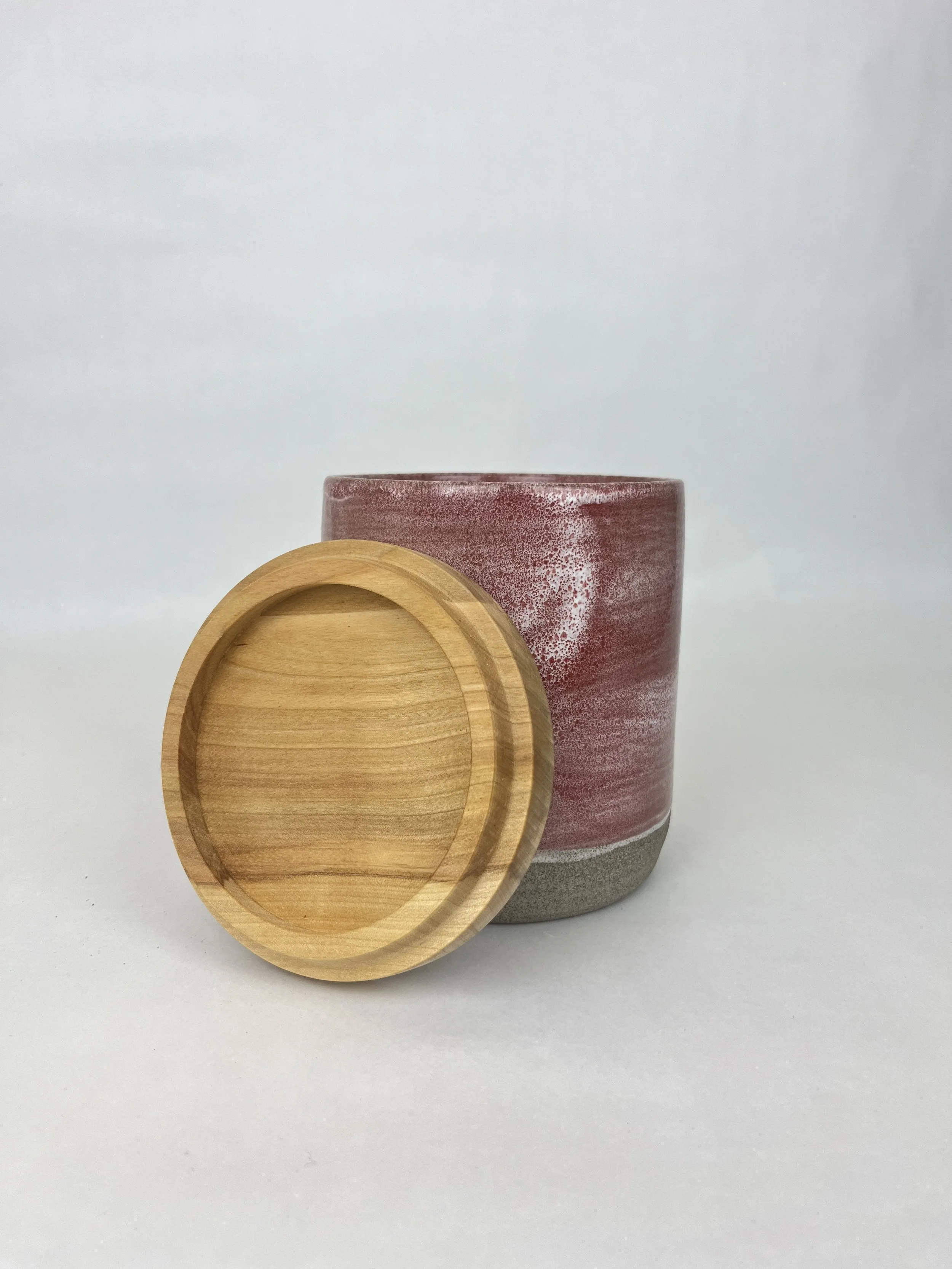 Ein roter Keramiktopf steht hinter einem runden Holzdeckel auf einem weißen Hintergrund.